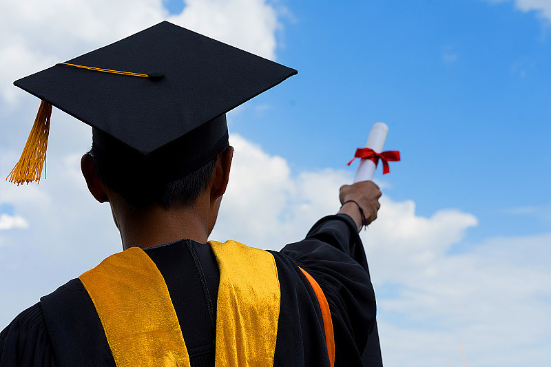 一名男性大学毕业生手持学位证书站在毕业典礼上庆祝，背景是湛蓝的天空。大学毕业生出示毕业证书。