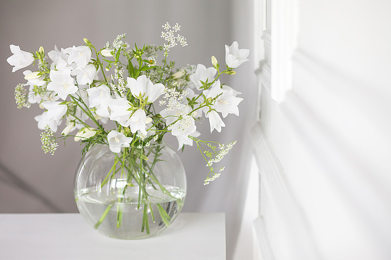 花瓶里一束轻柔的铃铛。房间里有晨光。柔软的家庭装饰，玻璃花瓶与白色的花在白色的墙壁背景和木制的桌子上。内部。贺卡。副本的空间。