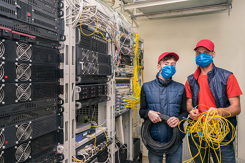 服务器室有两名带医用口罩的技术人员。专家手里拿着一卷电线，在数据中心的机架附近工作。服务工程师维护计算机设备