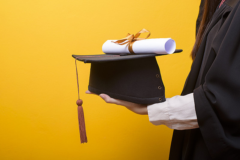 近距离的女子手在毕业礼服是拿毕业帽和证书的黄色背景