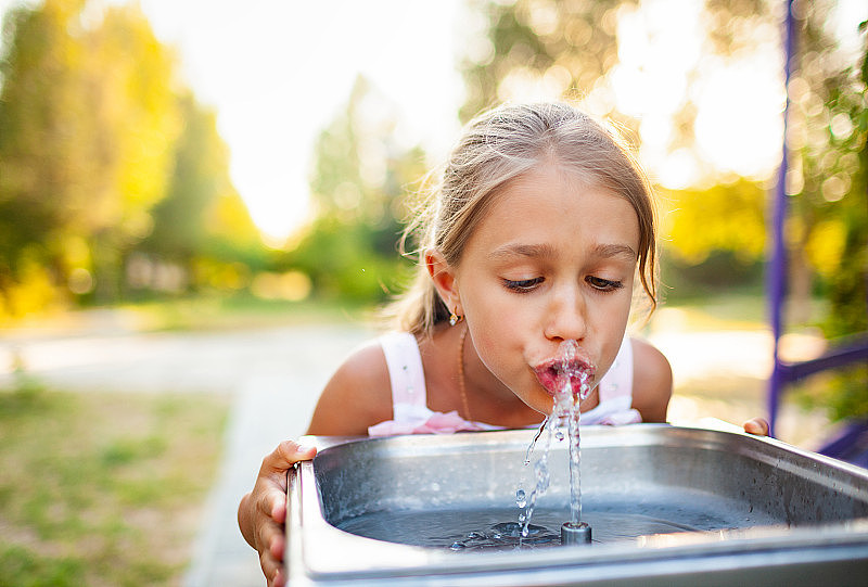 在一个夏日温暖阳光明媚的公园里，一个快乐的奇妙的女孩从一个小喷泉里喝着新鲜的水