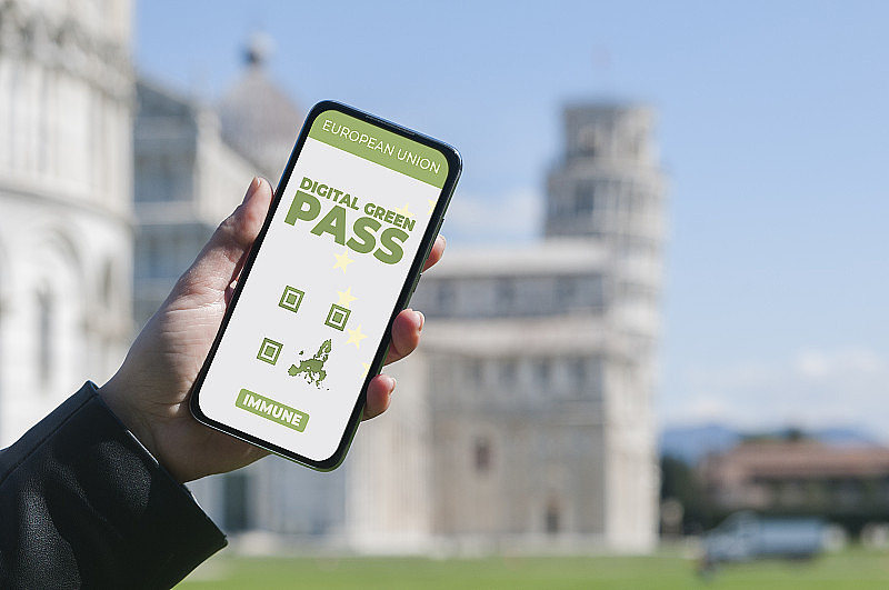 数字绿通行证的概念:一名女子在比萨斜塔前拿着智能手机，屏幕上显示着一个假想的数字绿通行证(或数字通行证证书)应用程序。
