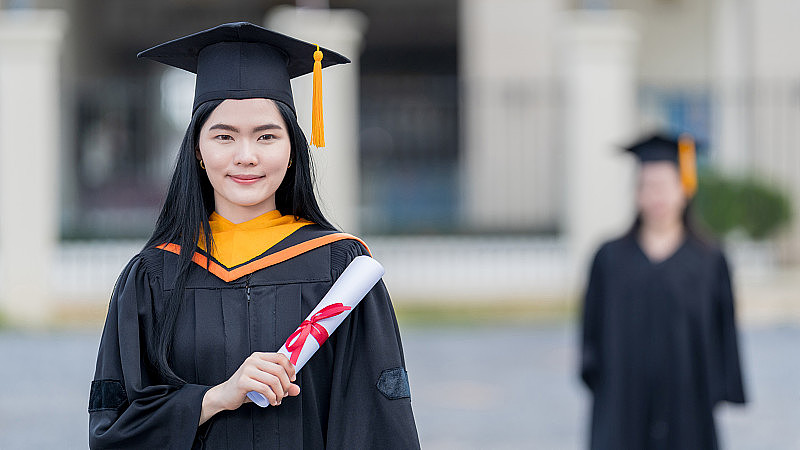 一位年轻美丽的亚洲女子大学毕业生穿着毕业礼服和学士帽，在参加大学毕业典礼后，手持学位证书站在大学大楼前