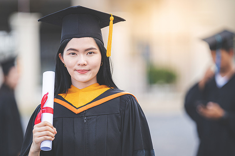 一位年轻的亚洲女子大学毕业生穿着毕业礼服和学士帽，在参加大学毕业典礼后，手持学位证书站在大学大楼前