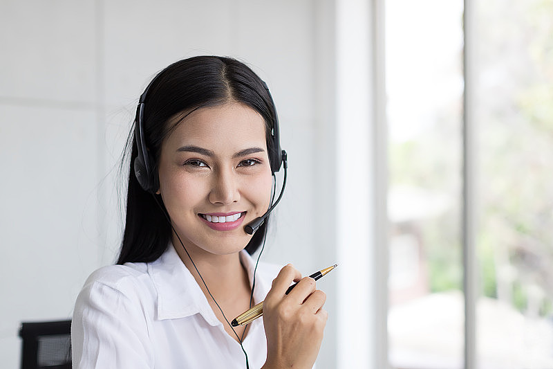 亚洲呼叫中心女工作人员。微笑的女客服接线员戴着耳机在办公室工作。呼叫中心和客户服务的耳机概念