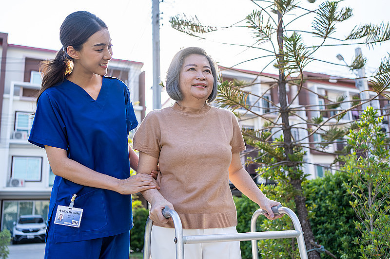 亚裔护理人员支持残疾妇女在户外行走。美丽的女孩医生帮助和照顾老年人成熟的残疾病人做物理治疗在公园的疗养院。
