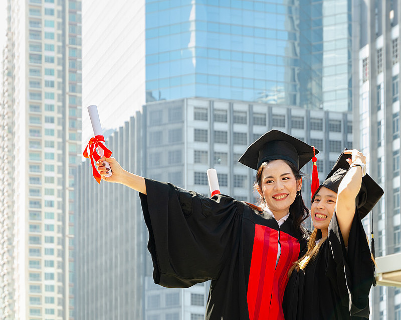 两个幸福的毕业女大学生，穿着黑色的毕业礼服，手拿毕业证，在毕业典礼上相互拥抱，站在城市建筑背景的户外