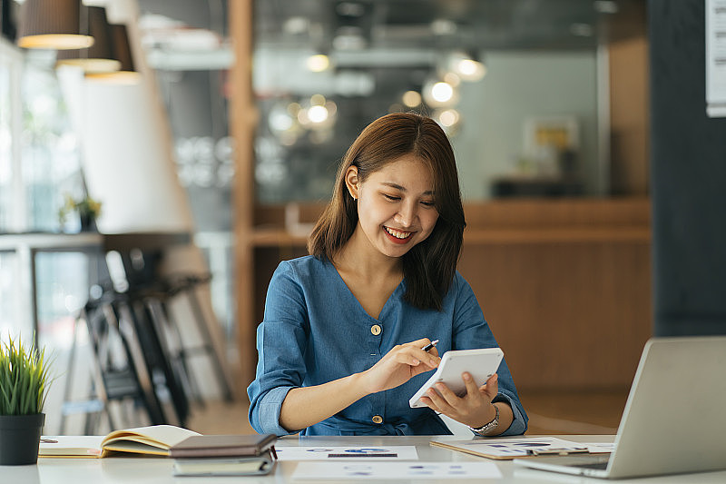 亚洲商业女性的肖像在办公室用笔记本电脑在线支付账单。有电脑和支票簿的漂亮女孩，乐意付账单。启动企业财务核算概念