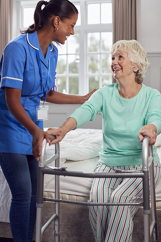 在家中使用步行架的老年妇女由穿着制服的女护理人员帮助下床