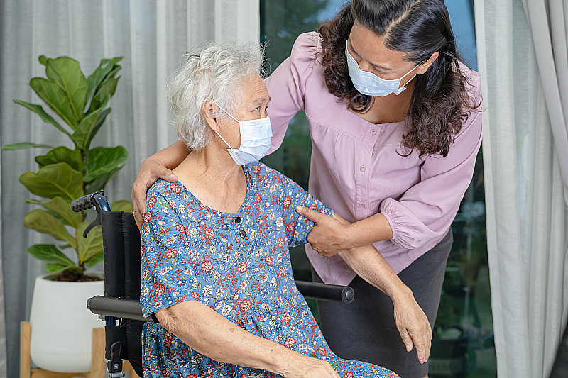 照顾者帮助亚洲老人或老人老妇人妇女坐在轮椅上，戴着口罩保护安全感染Covid - 19冠状病毒。