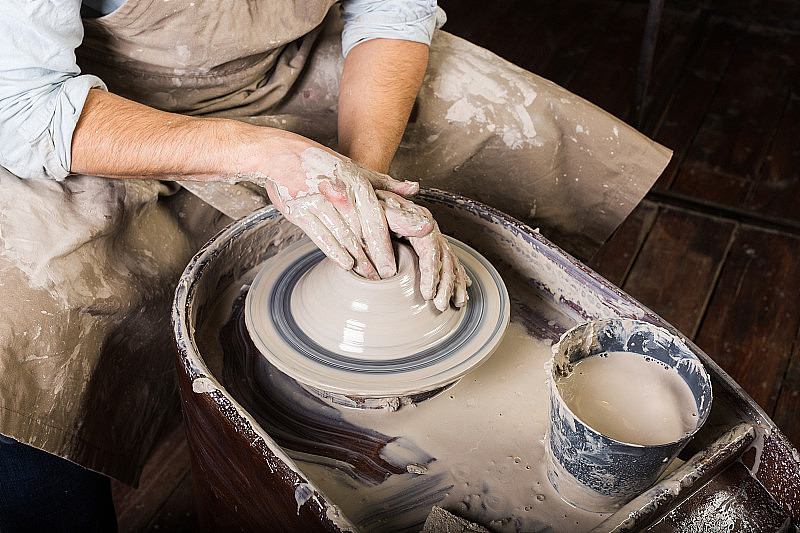 陶器，车间，陶瓷艺术概念-近距离工作的陶工车轮与原料粘土和人的手，一个男性雕刻器具与手指，在围裙，顶部视图