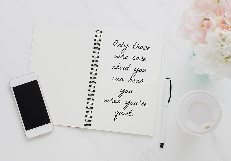 在笔记本上的智能手机，咖啡杯和白色大理石桌子上的花的鼓舞人心的报价。