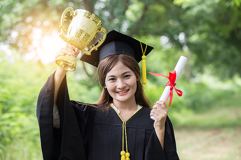 毕业典礼上，亚洲女性毕业生举起双手，手拿证书和奖杯庆祝