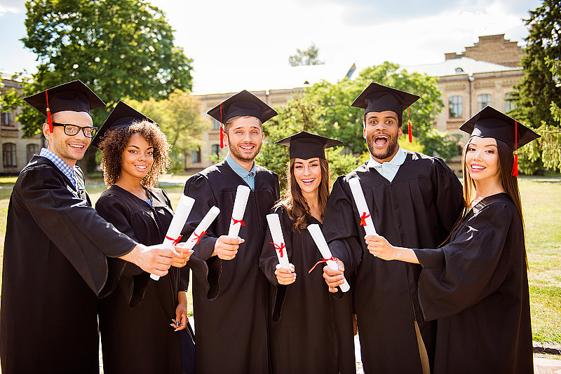 六名成功迷人的年轻毕业生身着黑色长袍，戴着帽子完成了他们的学业，手里拿着毕业证书，面带微笑，身后是学校大楼，阳光明媚